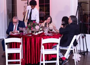 Dinner guests seated at tables with red tablecloths while being served by wait staff in formal setting.