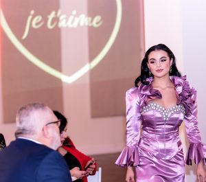 Fashion show model in lilac satin dress poses with 'je t'aime' heart projection in background.