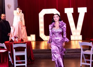Model in purple satin gown walks runway in front of illuminated LOVE letters at formal event.