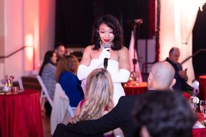 Speaker addresses dinner guests at formal event with red tablecloths and ambient lighting.