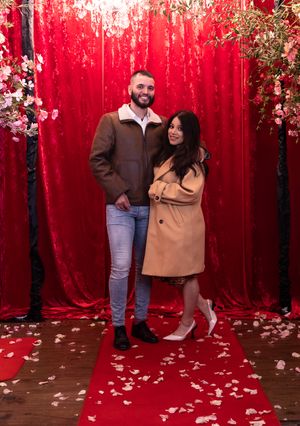 Couple in winter coats poses against a red velvet backdrop with cherry blossoms and scattered petals.