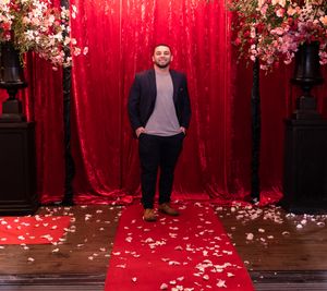 Man in navy blazer poses on red carpet against velvet backdrop with floral decorations.