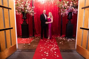 Couple poses in front of a red backdrop with floral decorations and scattered petals on the floor.