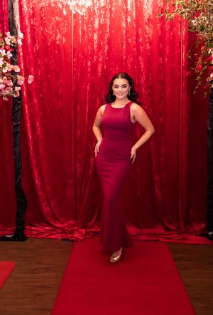 Figure in red gown poses gracefully against luxurious red velvet backdrop with floral accents.