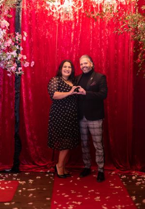 A couple poses together making heart shapes against a red velvet backdrop with floral arrangements.