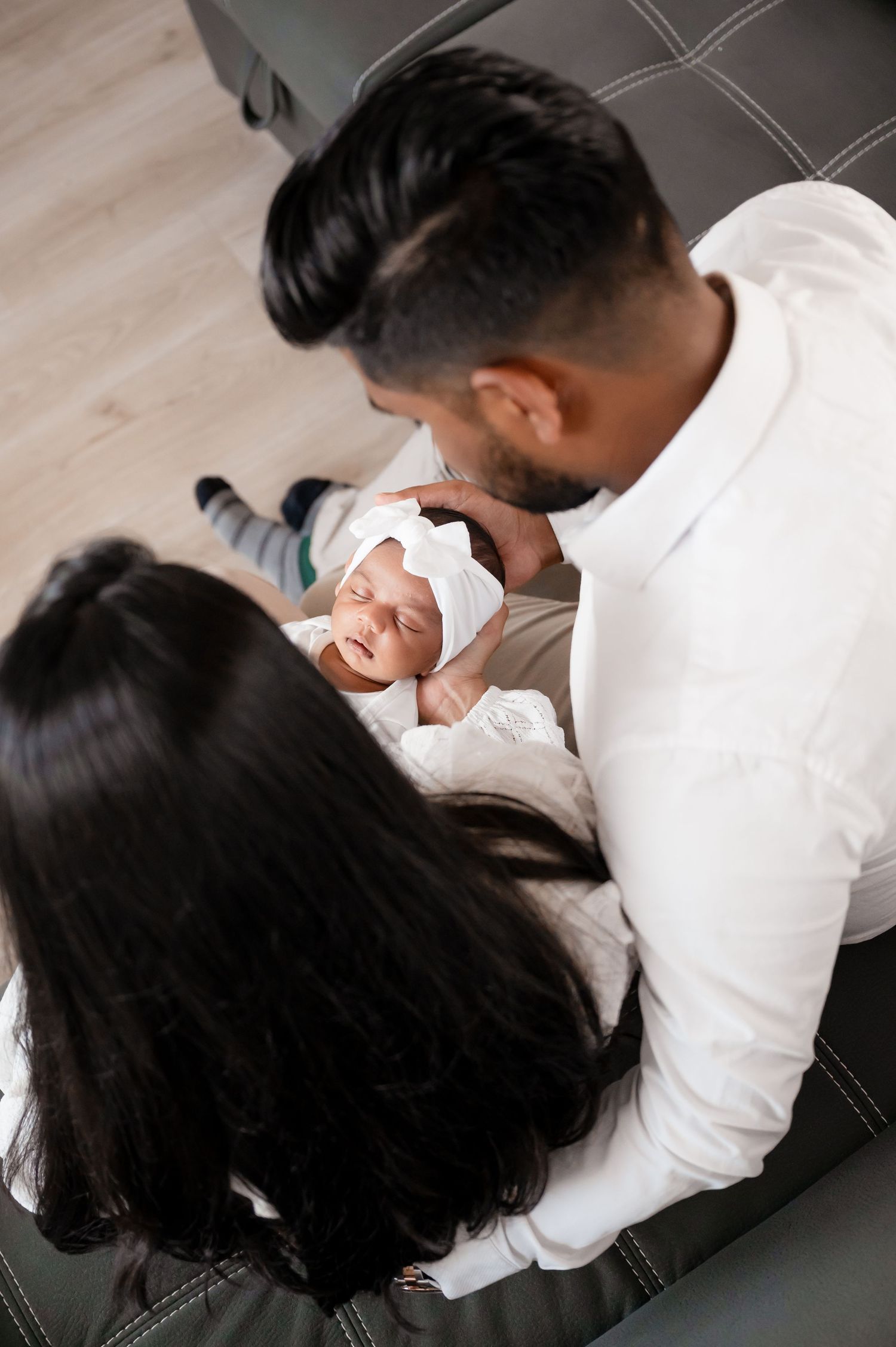 Parents in white clothing share a joyful moment with their smiling baby on a leather couch.