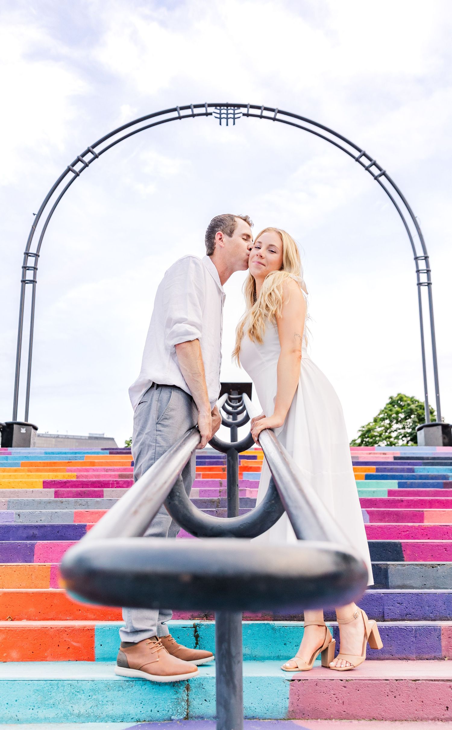 Couple poses on colorful rainbow stairs under a metal arch structure.
