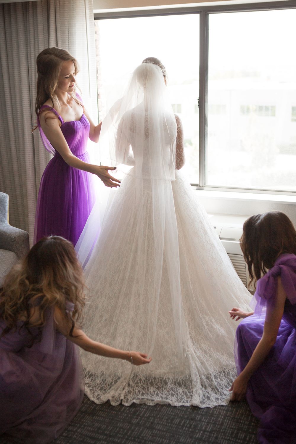 Wedding dress with lace details being arranged by bridesmaids in purple dresses near sunlit windows.