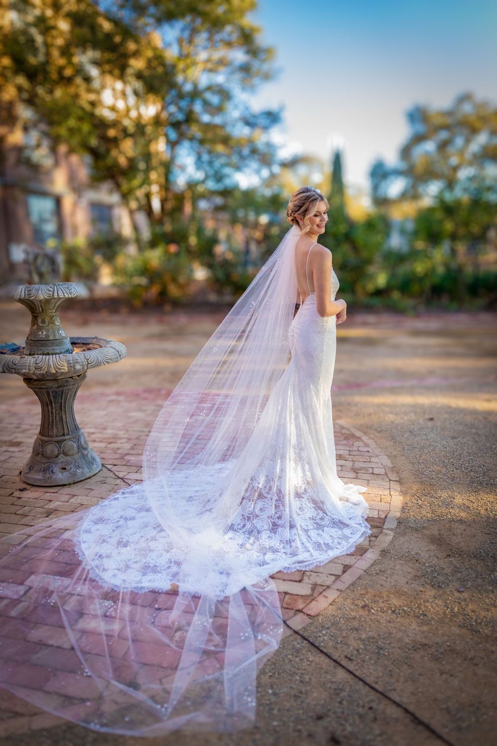 A flowing cathedral-length veil trails behind on rustic stone pavement in golden afternoon light.