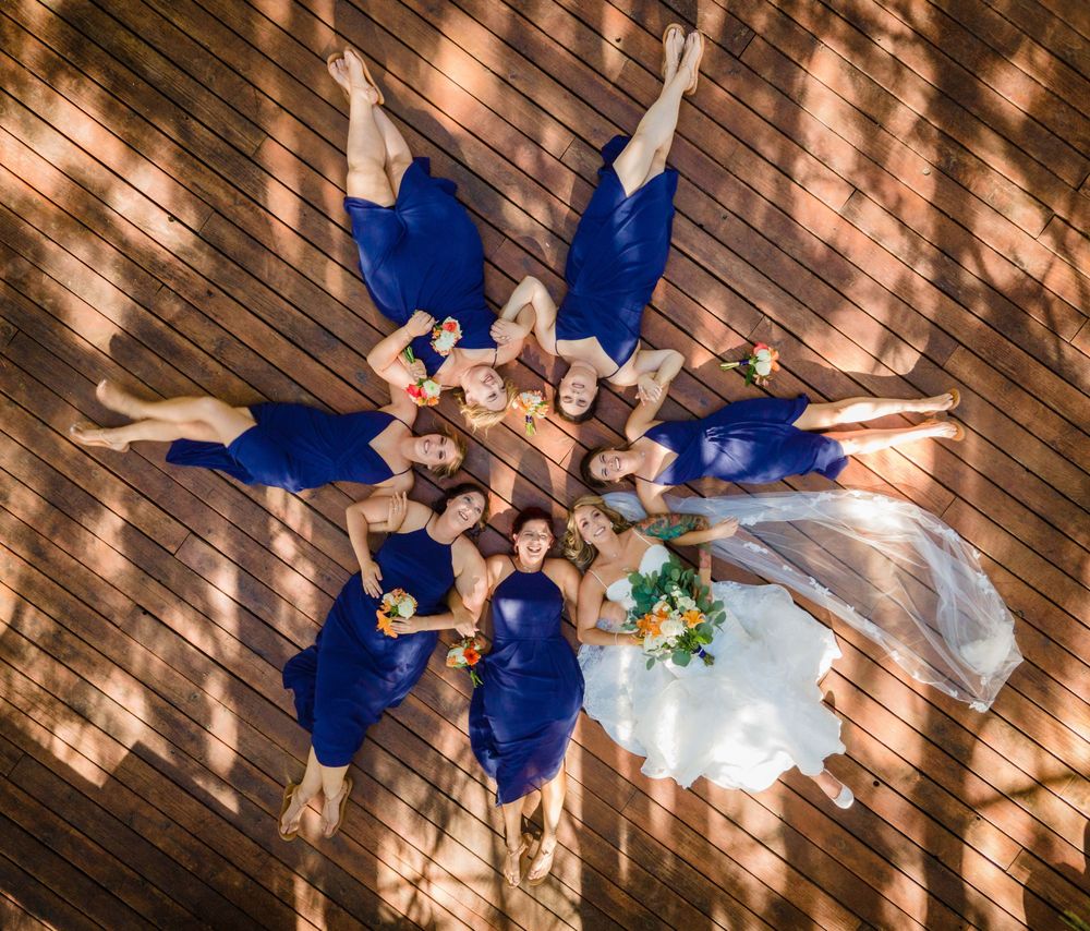 Aerial view of wedding party in royal blue attire lying on wooden deck in dappled sunlight.