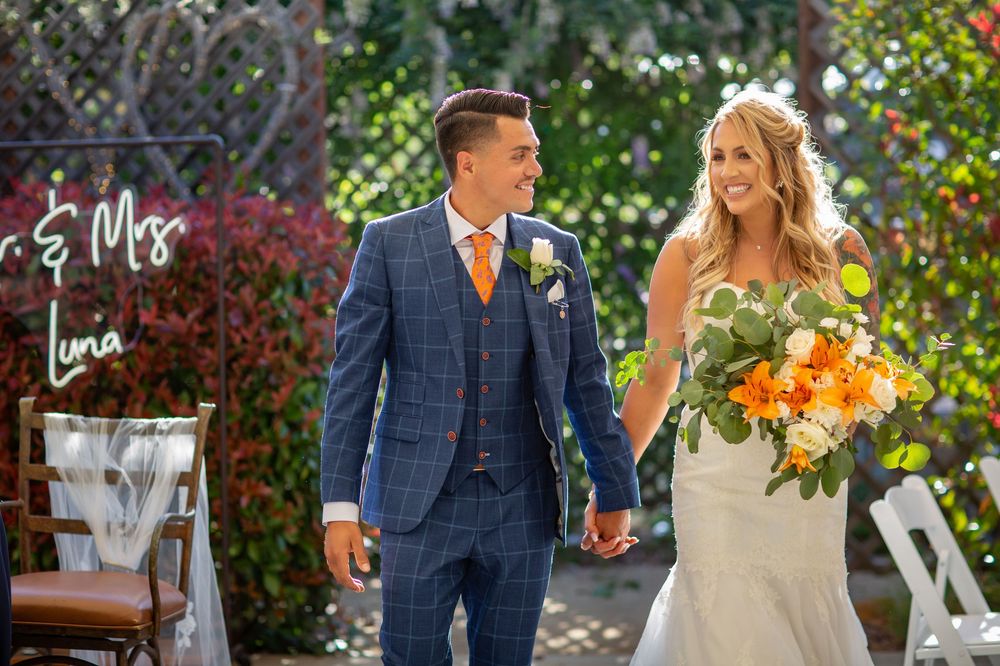 Wedding couple walks hand in hand through a lush garden setting with vibrant orange floral bouquet details.