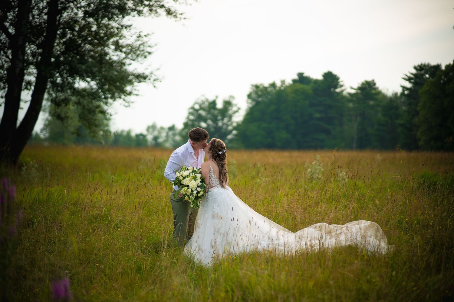 A bride and groom share a kiss in a field, the bride’s long train spread out behind her as they stand among tall grass.