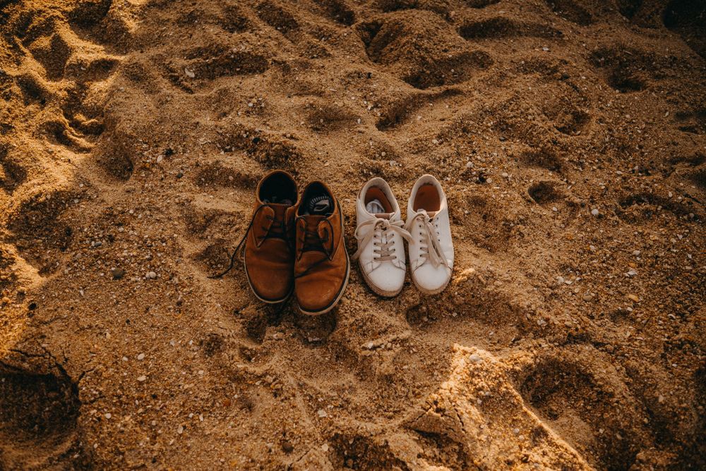 Two pairs of shoes, one brown and one white, sitting on sandy beach ground.
