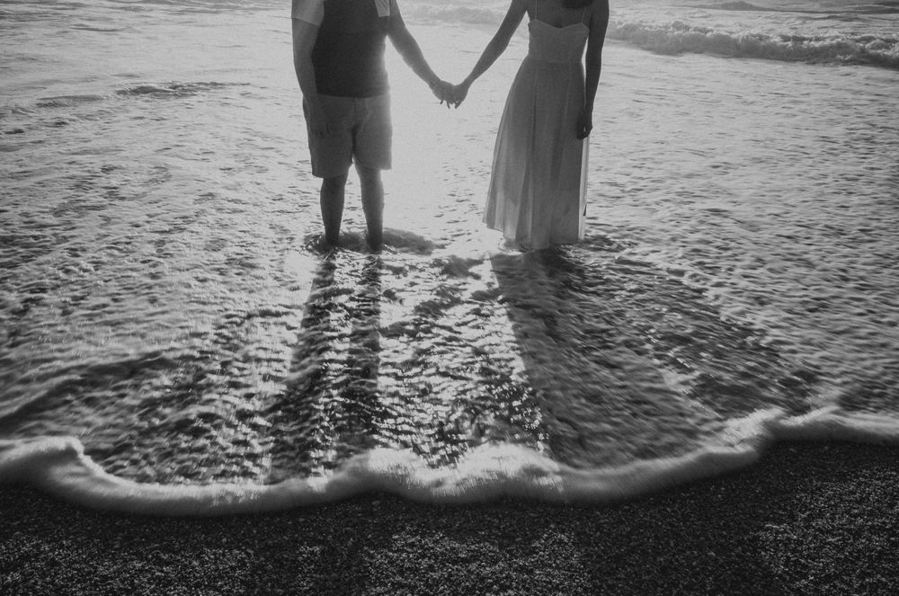 A silhouetted couple holding hands as ocean waves wash over their feet at sunset.