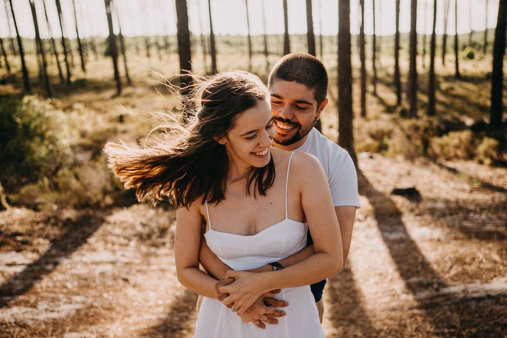 Couple laughing and embracing in a sun-dappled forest setting.