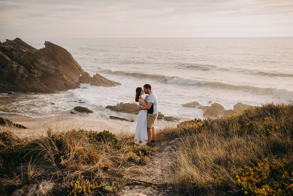 Couple standing on coastal cliffs overlooking dramatic ocean waves at sunset.