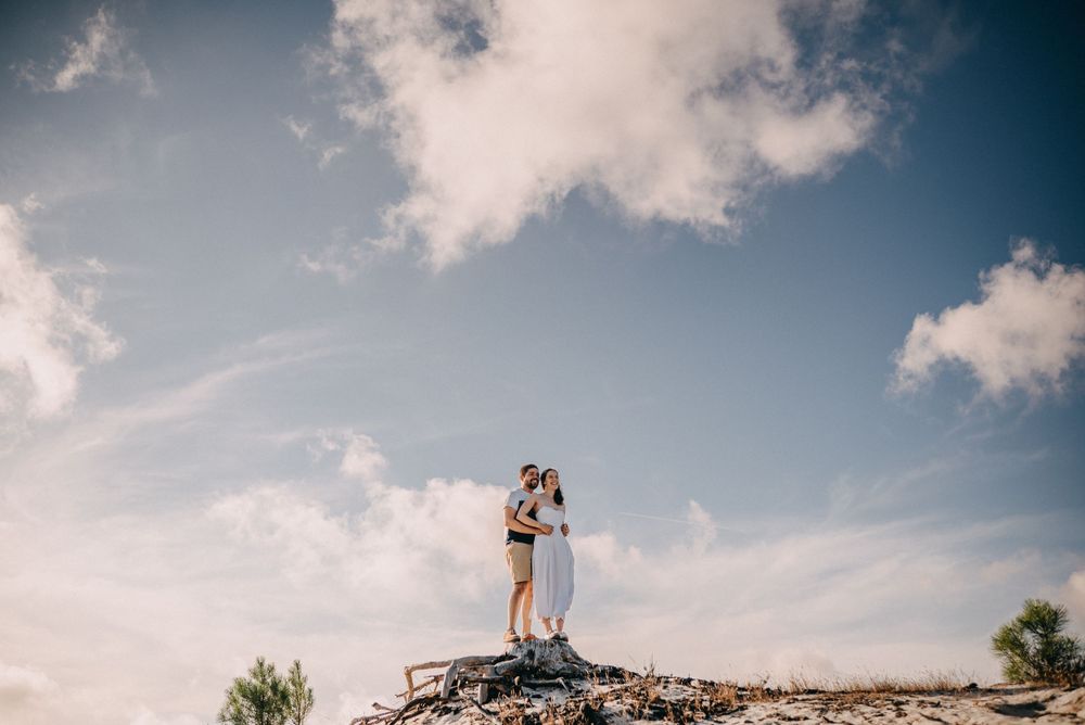 Couple embracing on a hilltop against a bright blue sky with fluffy clouds.