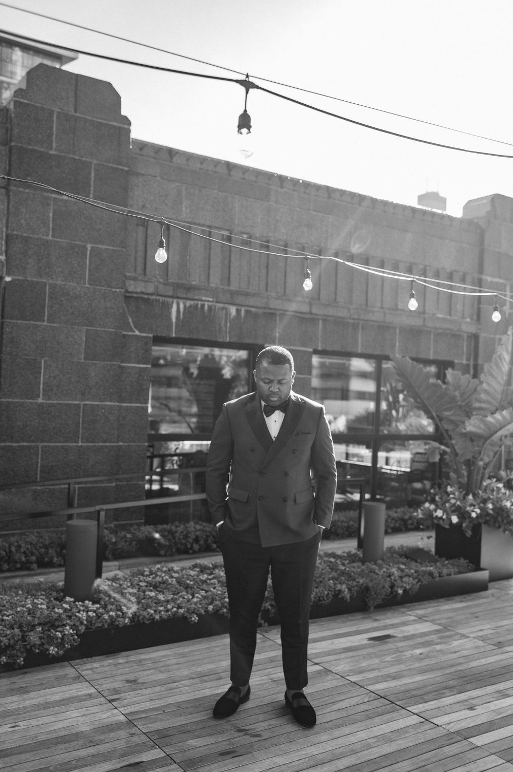 Person in formal attire standing on urban rooftop deck with string lights.