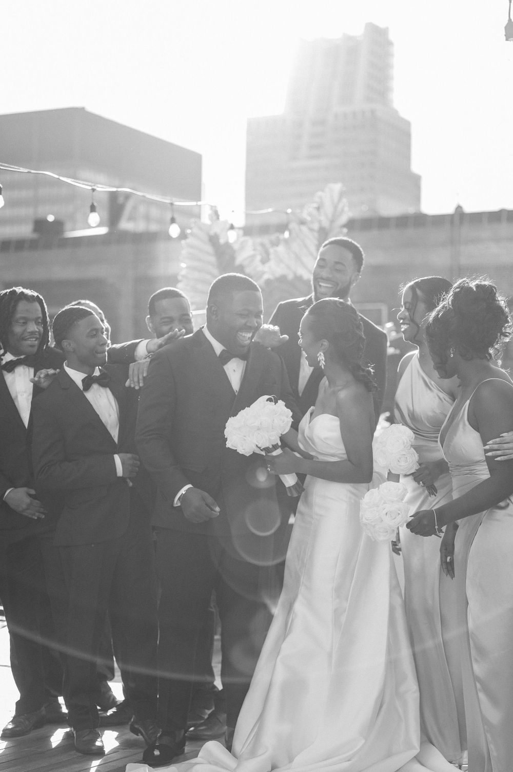 Wedding party gathered for group photo on sunny rooftop with cityscape background.