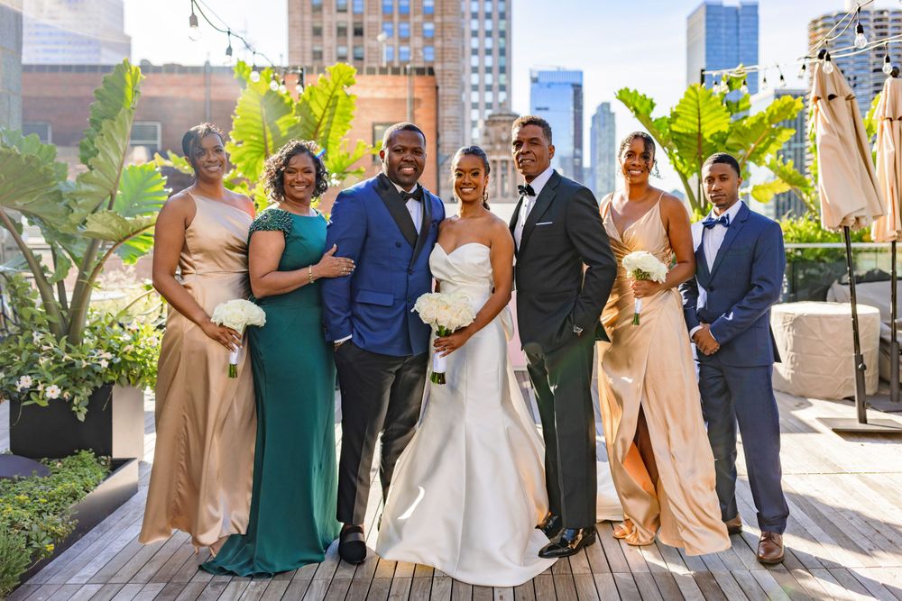 Wedding party poses outdoors on rooftop venue with urban cityscape and tropical plants in background.