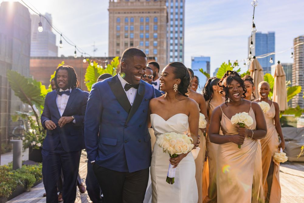 Wedding party walks together outdoors with city buildings and greenery in late afternoon sunlight.