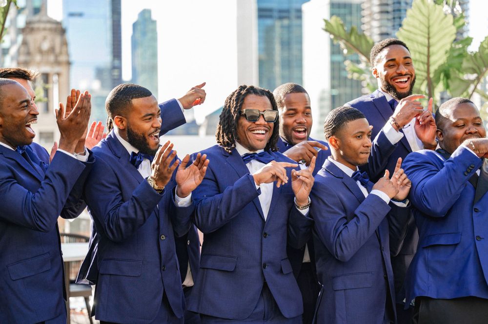 Groomsmen in matching navy suits pose for photos outdoors with city skyline in background.