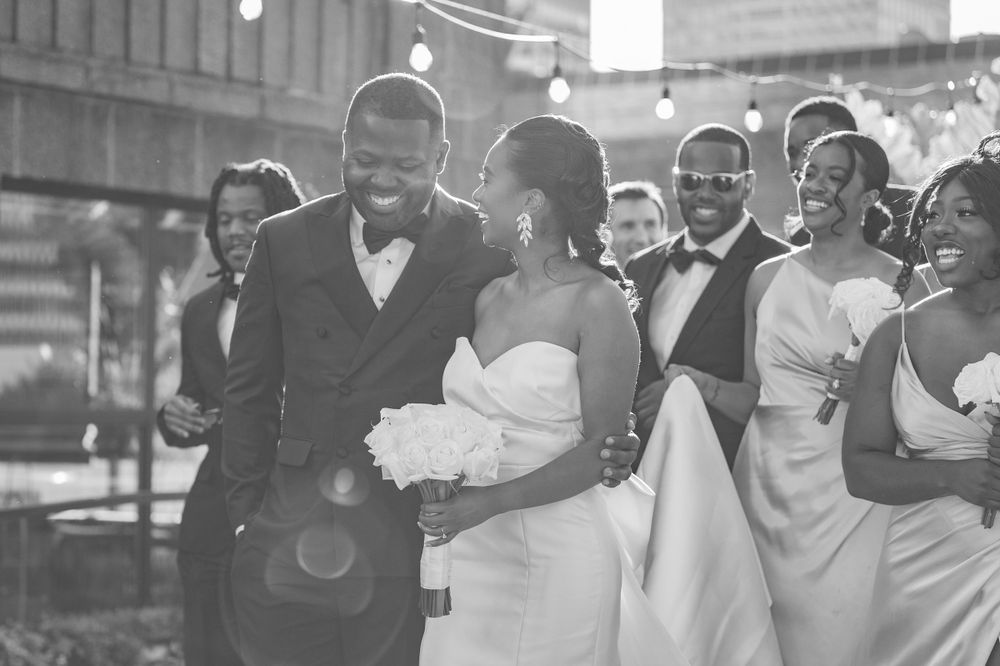 Candid black and white photo captures wedding party laughing together during outdoor celebration.