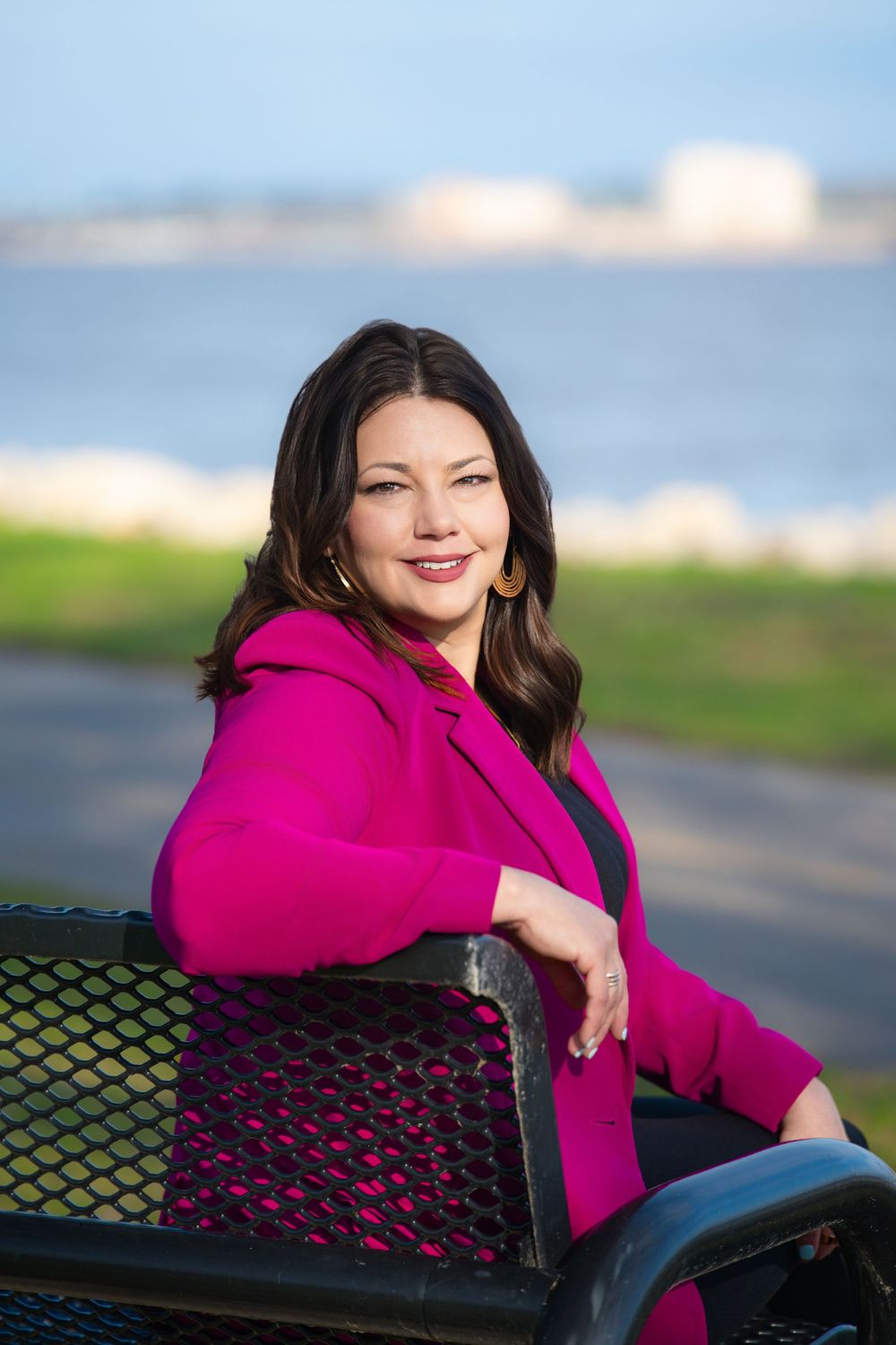 Woman in magenta blazer sitting on park bench with waterfront view smiling at camera.