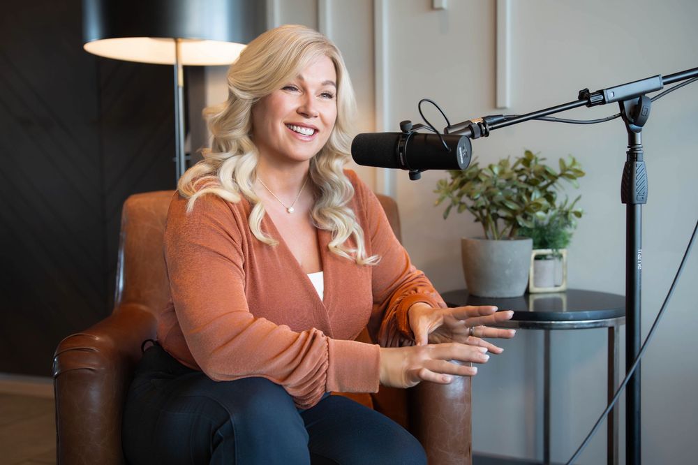 Woman with blonde hair in orange cardigan sits in leather chair speaking into podcast microphone in modern studio.