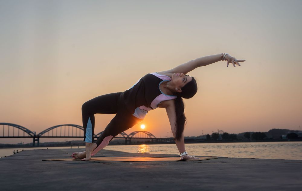 Woman practicing yoga in a triangle pose at sunset by a river with a bridge silhouetted in the background. Quad Cities Photographer