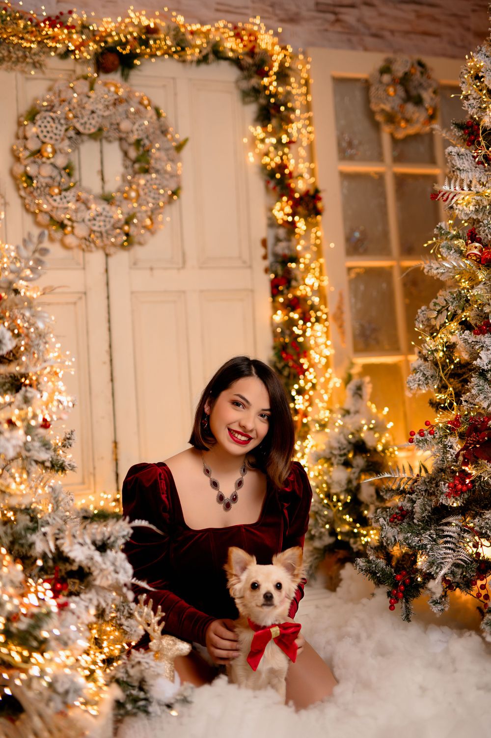 Elegant holiday portrait with a small dog in a red bow against a backdrop of twinkling lights and Christmas decorations.