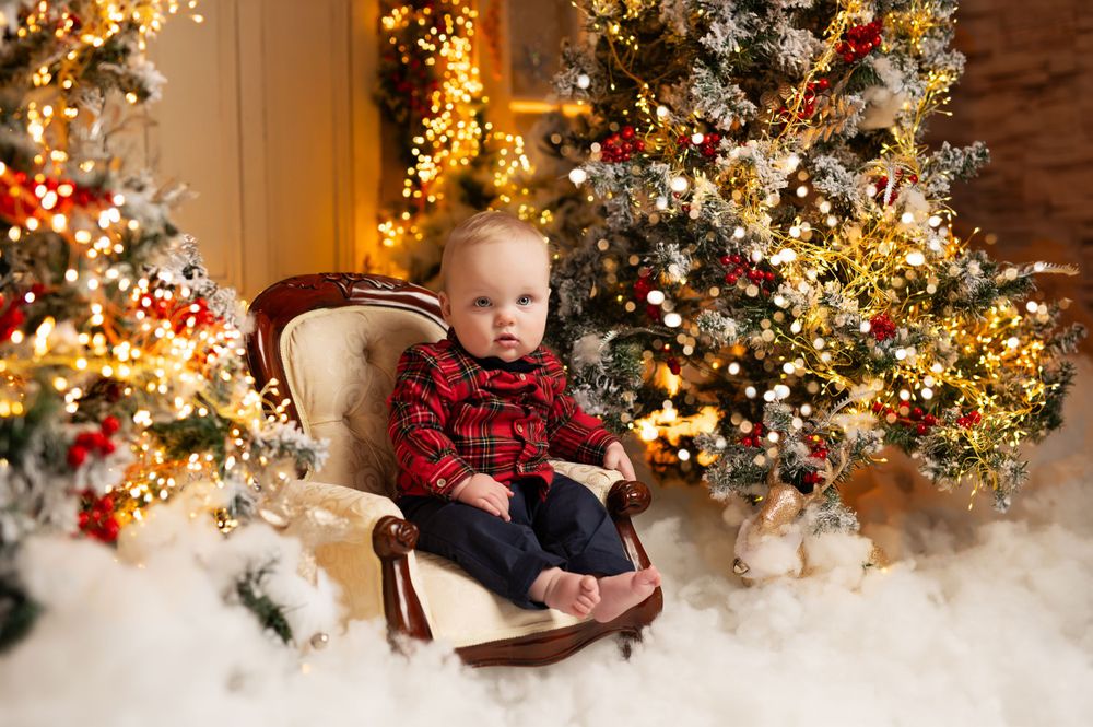 Baby in plaid sitting on vintage chair surrounded by glowing Christmas trees and holiday decorations.