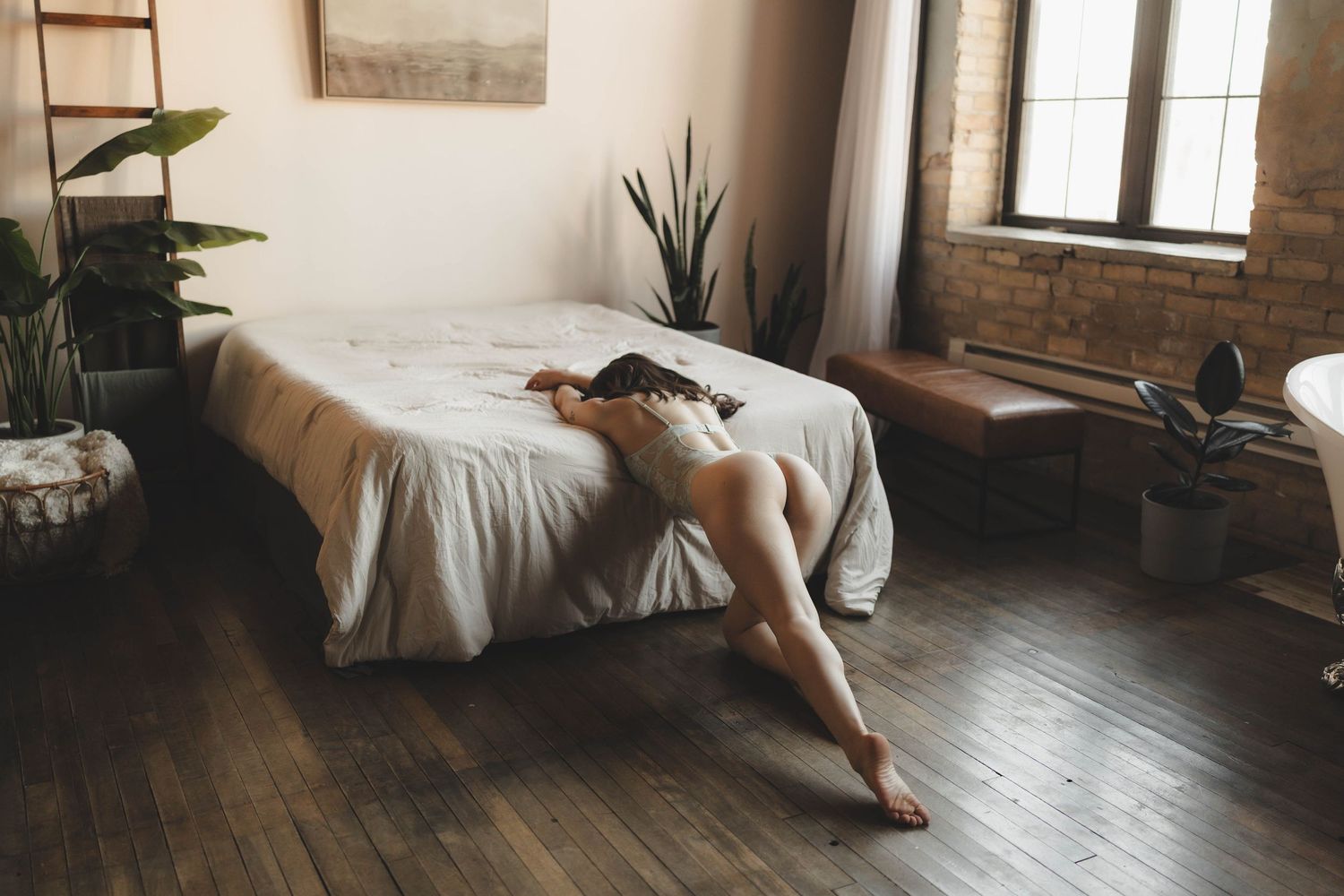 A woman in a gray off-shoulder top poses gracefully on a hardwood floor beside a bed in a modern bedroom.