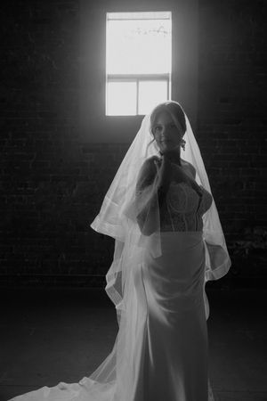 Black and white bridal portrait of a bride in a wedding gown and veil, backlit by a window in a dark brick room.