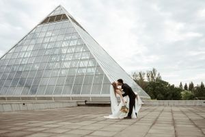 A wedding couple poses in front of a modern glass pyramid architectural structure on a cloudy day.