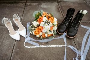 Wedding accessories including white heels, black boots, and orange and white floral bouquet arranged on stone floor with ribbons.