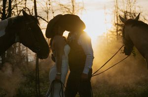 Couple in cowboy hats sharing a kiss at golden sunset between two horses in a romantic western wedding scene.