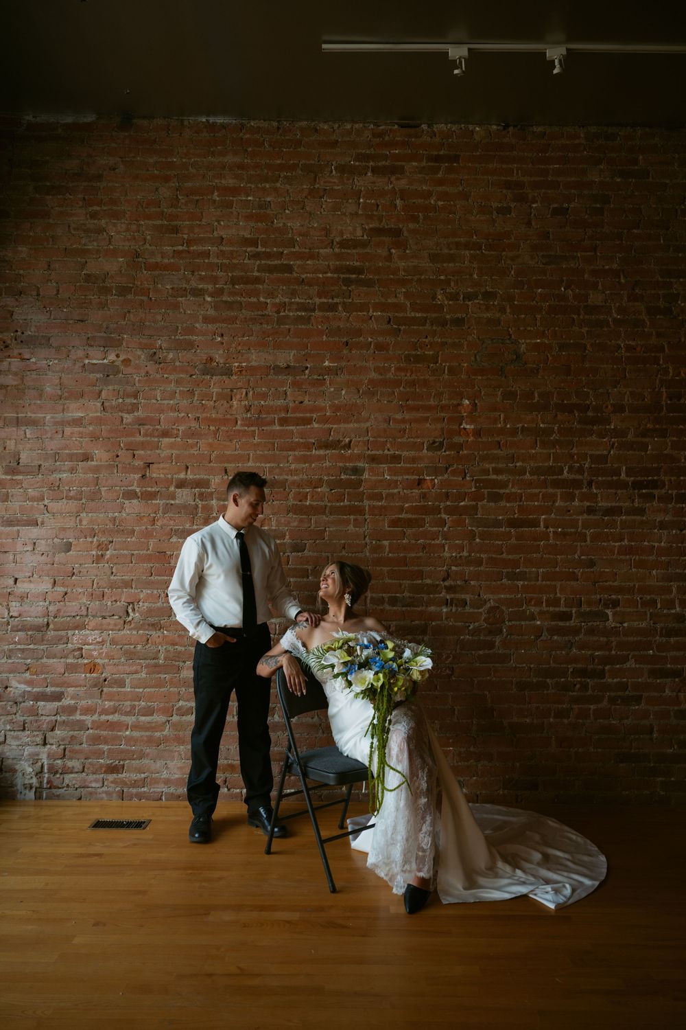 Bride seated on chair holding blue floral bouquet with groom standing beside her against rustic brick wall on wood floor.