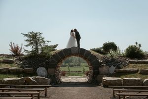 A silhouetted couple stands atop a rustic stone bridge archway in a garden setting at sunset.