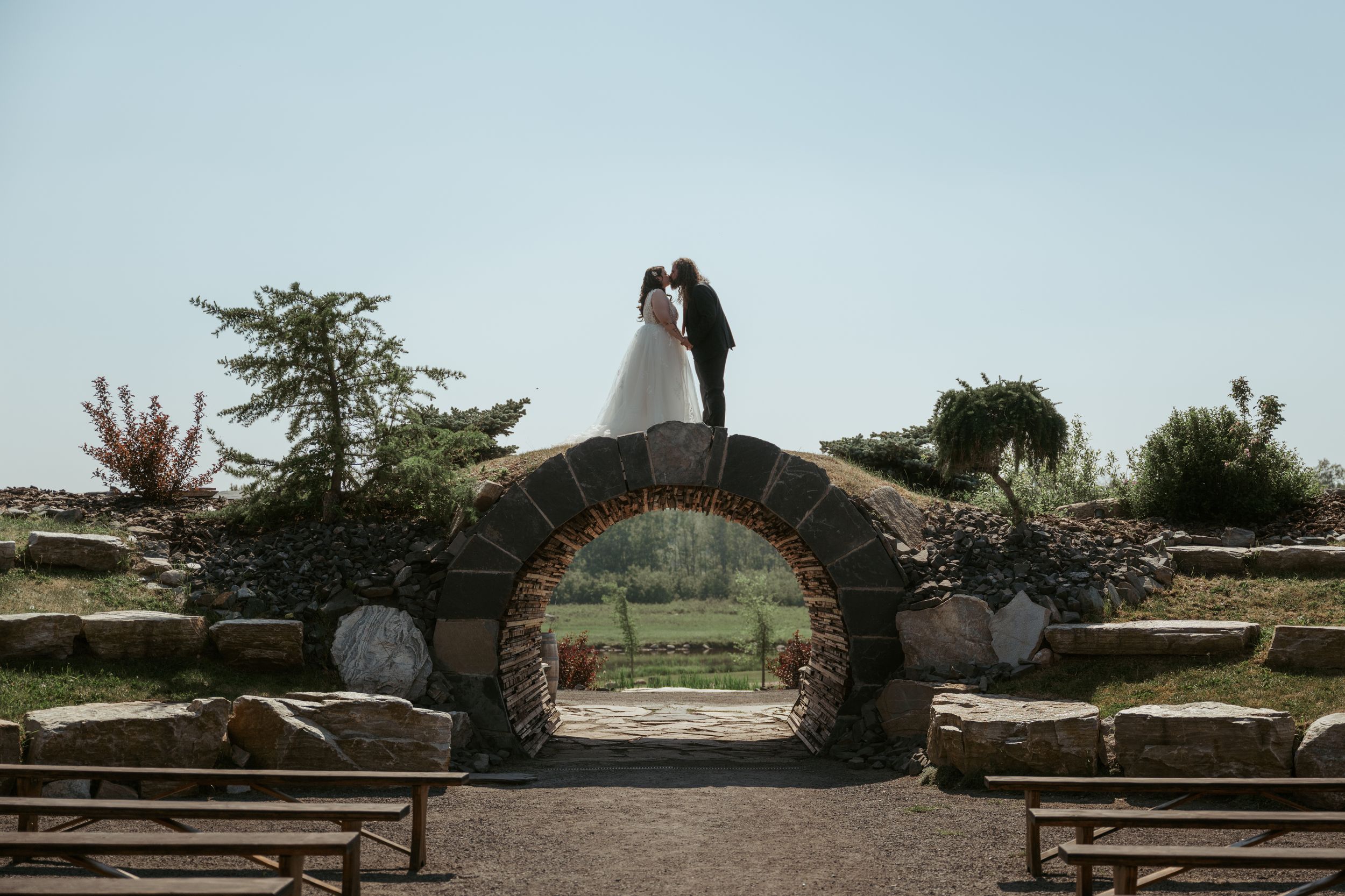 A silhouetted couple stands atop a rustic stone bridge archway in a garden setting at sunset.