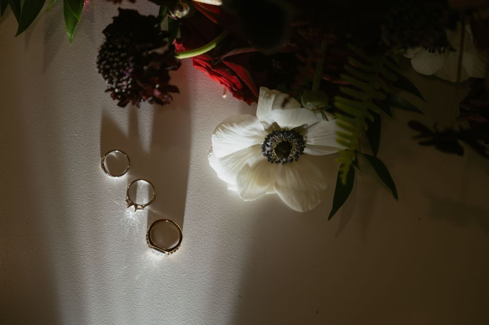White anemone flower with gold hoop earrings and locket on white surface with dark floral bouquet in moody wedding flat lay.