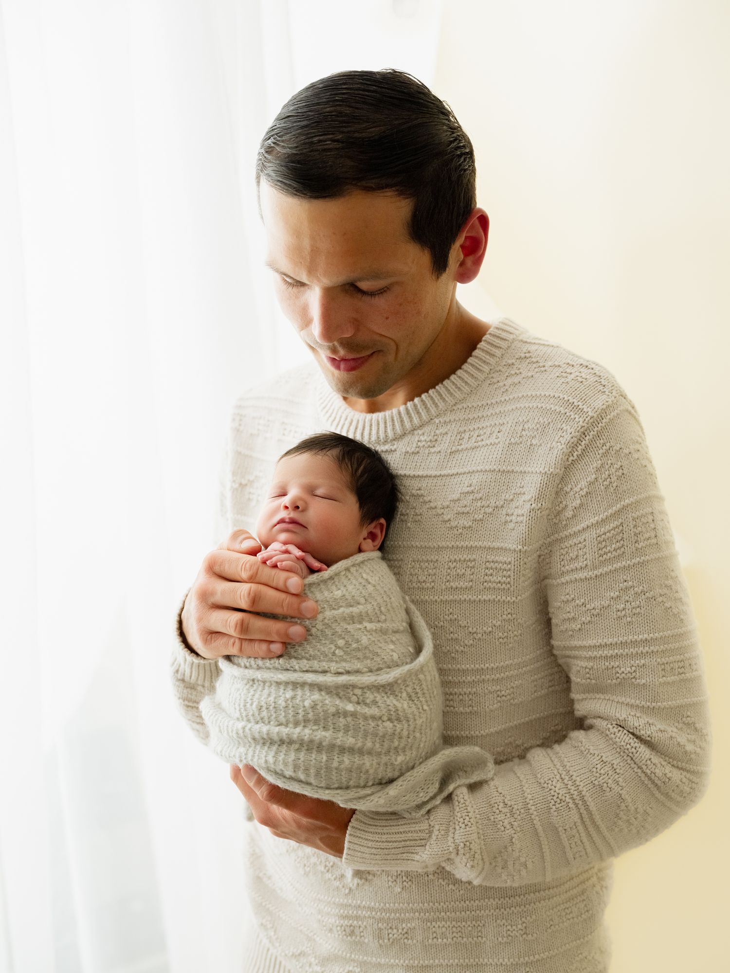 Parent in cream cable knit sweater tenderly holds and gazes at sleeping newborn baby in minimalist white setting.