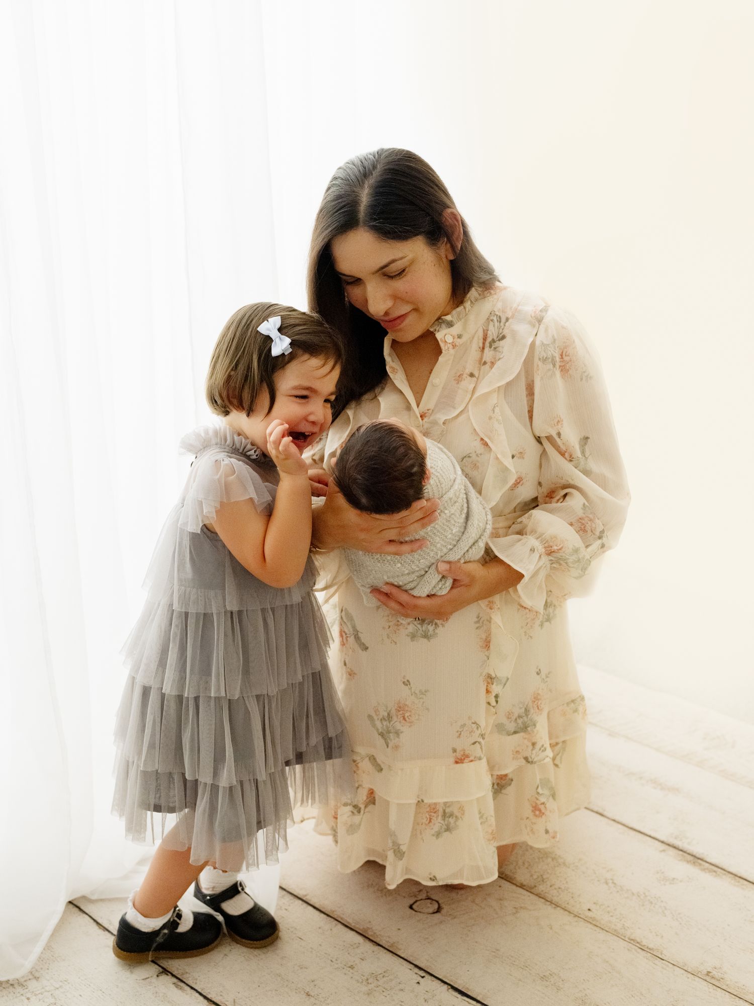 A mother in a floral dress and daughter in a gray tutu dress share tender moments in a bright white room.