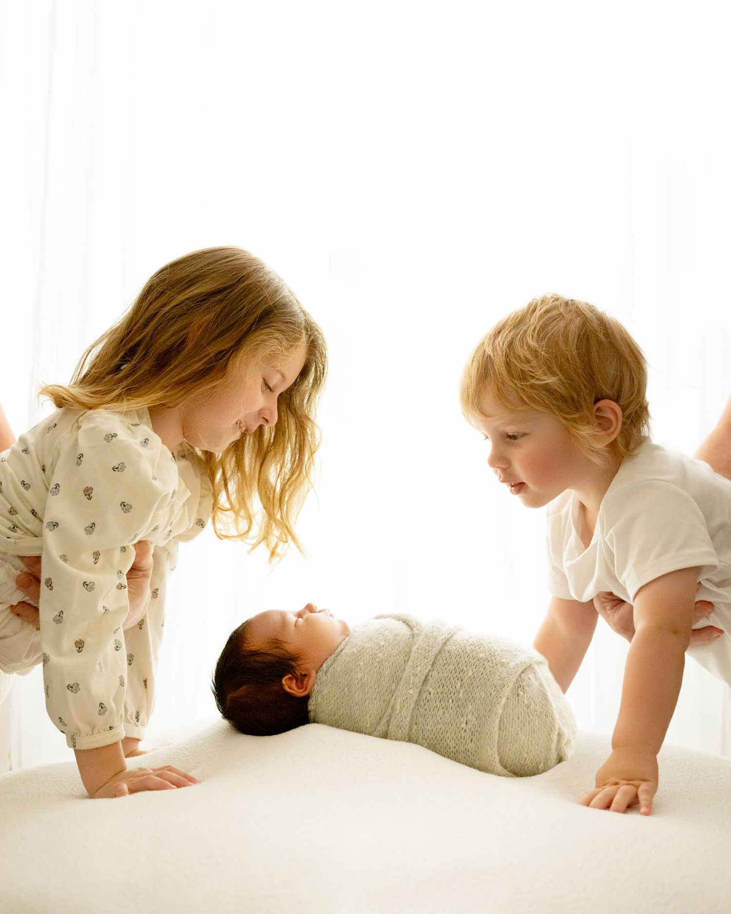 Two young children look down at a swaddled newborn baby laying on a white surface in bright natural light.