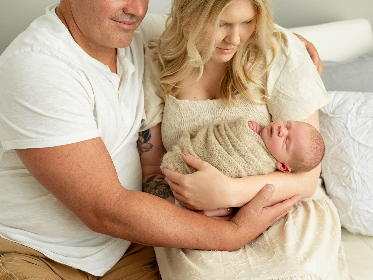 Parents in white clothing holding their new infant wrapped in beige blanket on bed with a white background.