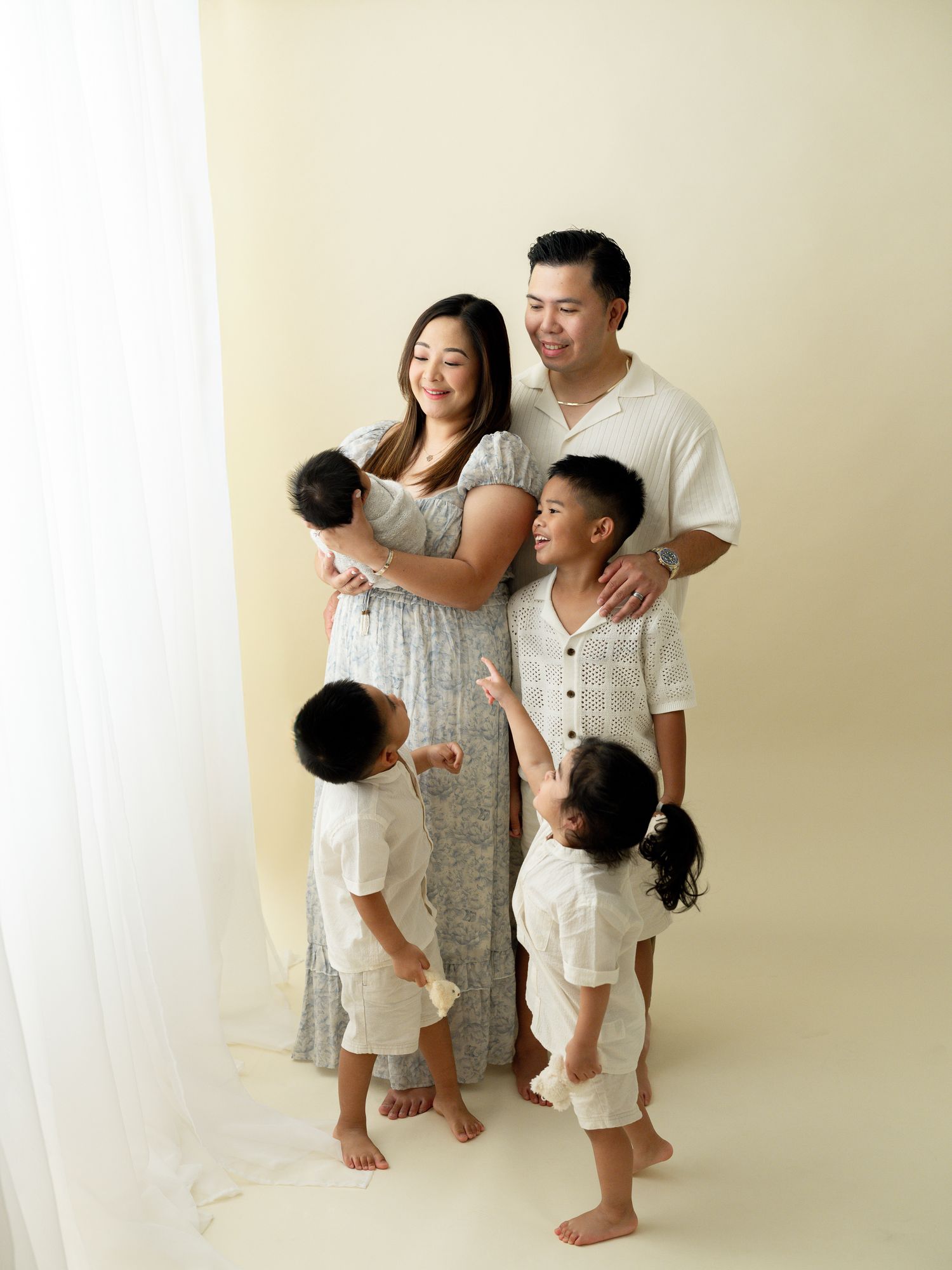 Family wearing coordinated white and cream outfits poses together against beige backdrop in studio portrait.