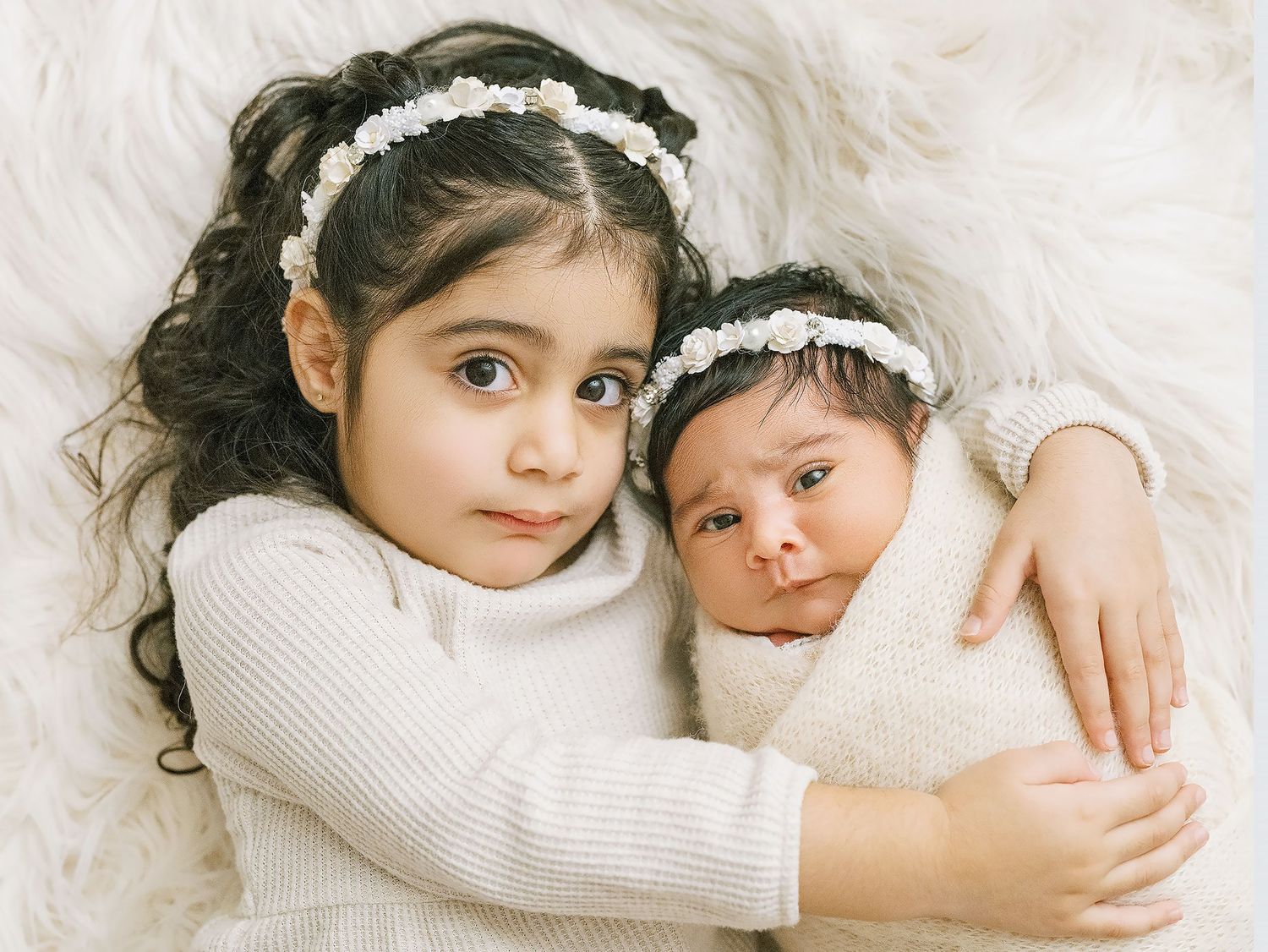 Two children wearing white headbands and sweaters embrace while lying on a soft furry background.
