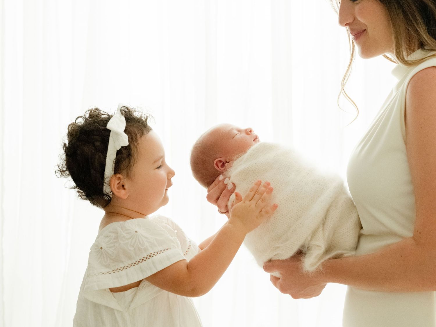 A baby in white clothing sleeps peacefully while being held in gentle hands against a bright window background.
