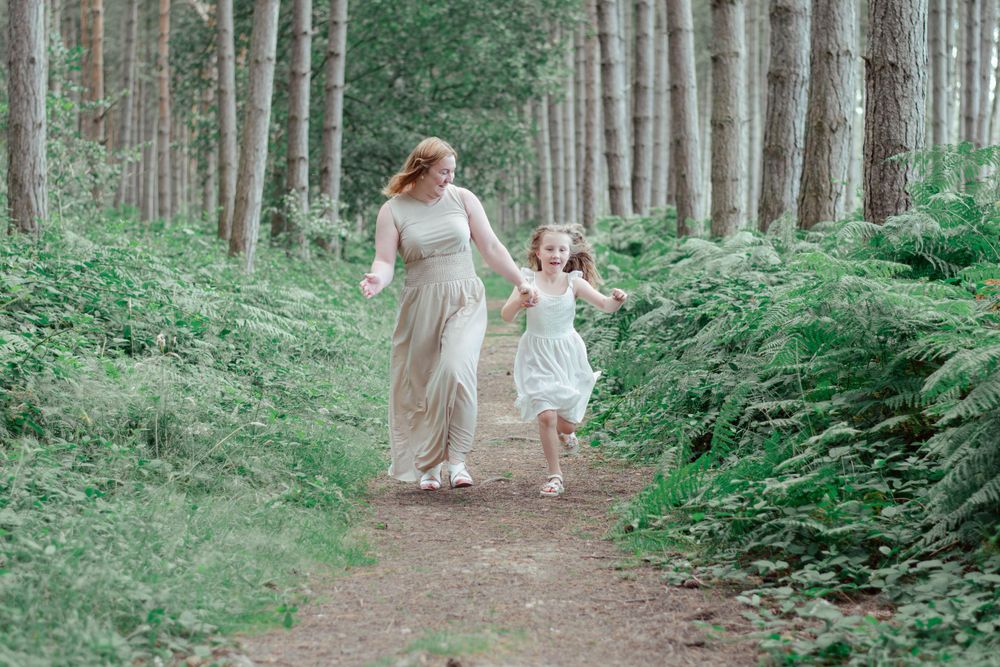 Mother and child in white dresses running together along a forest path surrounded by tall trees.