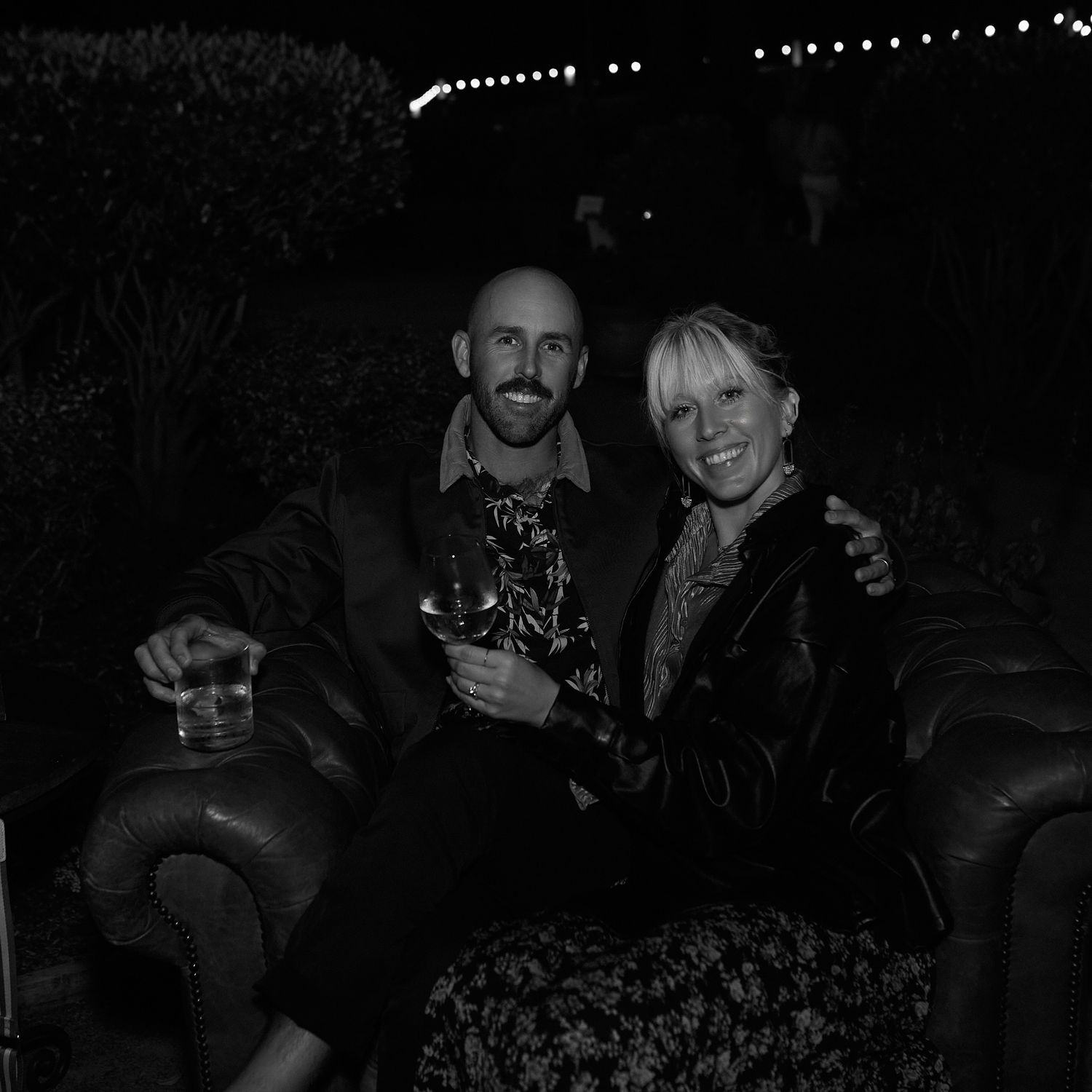 Black and white nighttime photo of two people sitting together on a couch under string lights.